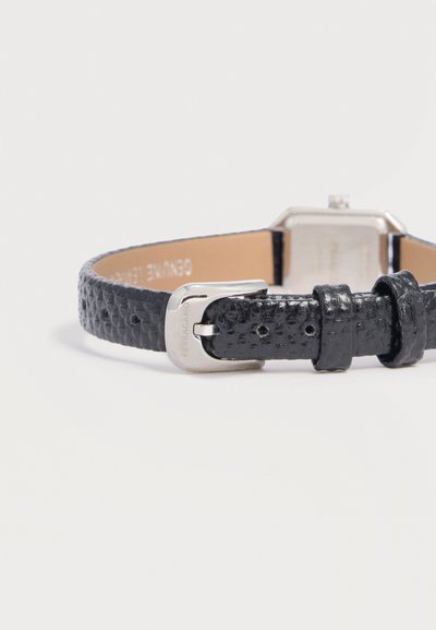 Bracelet de montre en cuir noir texturé avec boucle argentée embossée "Ferragamo", et un boîtier de montre argenté flou sur une surface blanche.