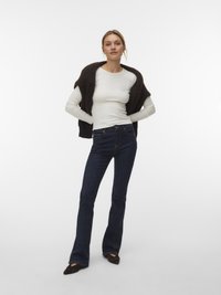 Frau trägt dunkelblaue Schlagjeans, weißes langärmliges Oberteil, dunklen Strickpullover über die Schultern gelegt und dunkle spitze Schuhe.