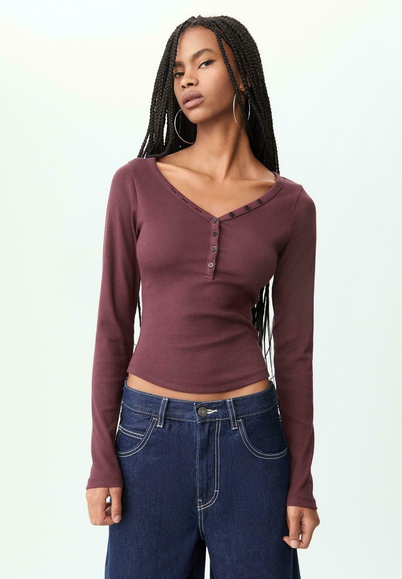 Long-sleeve, burgundy ribbed top with a V-neck and five-button detail at the neckline, paired with high-waisted blue denim jeans.