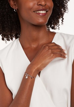 Zilveren charmearmband met een hangslot en accenten van een halve maan. Draagt op een hand met een elegante, gladde huidtextuur. Wit V-hals top.