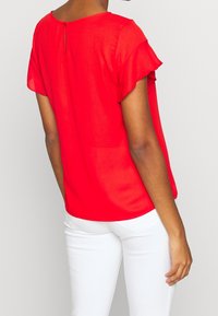 Blusa rossa a maniche corte realizzata in un tessuto leggero e morbido. Presenta un collo rotondo e una piccola apertura sul retro con chiusura a bottone.