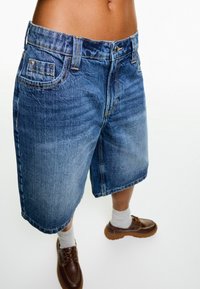 Jeansshorts i medelblå tvätt, med rak bendesign, standardfickor och kontrasterande sömdetaljer. Bärs med bruna skor och grå strumpor.