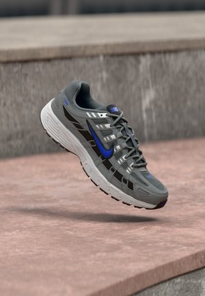Chaussure de course Nike grise et noire avec un swoosh bleu et une semelle blanche flottant au-dessus d'une surface en pierre.
