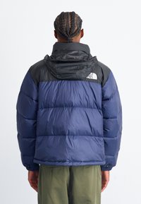 Mørkeblå stim-jakke med en sort, polstret hætte og skulderområde. Har en blank overflade og North Face-logo på ryggen.