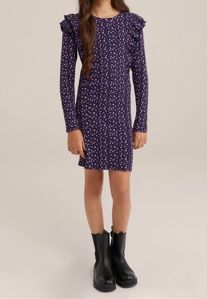 Fille portant une robe violette à manches longues avec de petites fleurs roses et blanches imprimées et des bottines noires, debout devant un fond uni.