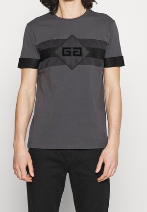 Mężczyzna ubrany w szarą koszulkę z krótkim rękawem, z czarnymi poziomymi paskami oraz czarnym geometrycznym emblematem na środku klatki piersiowej.