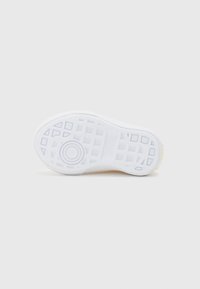 Semelle de chaussure en caoutchouc blanc avec un motif de grip texturé, emblème circulaire au centre et légère accentuation jaune près du talon.