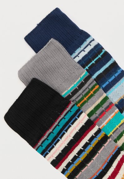 Trois chaussettes en tricot pliées en noir, gris et marine avec des rayures horizontales multicolores empilées sur un fond blanc.