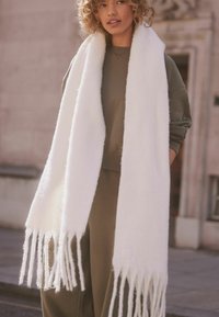 Weißer, flauschiger Schal mit langen Fransen, aus weichem Material, kombiniert mit einem grünen Sweatshirt und Hose, vor einem Outdoor-Hintergrund präsentiert.
