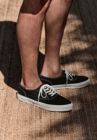 Scarpe da ginnastica nere in tela con lacci bianchi, design a profilo basso e suole in gomma. Lo sfondo presenta una superficie intrecciata e strutturata.