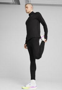 Zwart sporttop met lange mouwen en bijpassende snug-fit leggings, met een reflecterend logo. Het model draagt witte sneakers met neonaccenten.