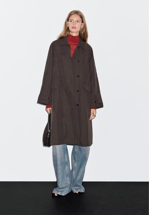 Lang, mørkebrun frakke lavet af stof, med knapper, sidelommer og en afslappet pasform, parret med lyseblå brede jeans.