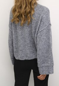 Grå stickad tröja med mjuk textur, kort design, breda ärmar och ribbade detaljer vid muddarna. Bärs med svarta byxor.