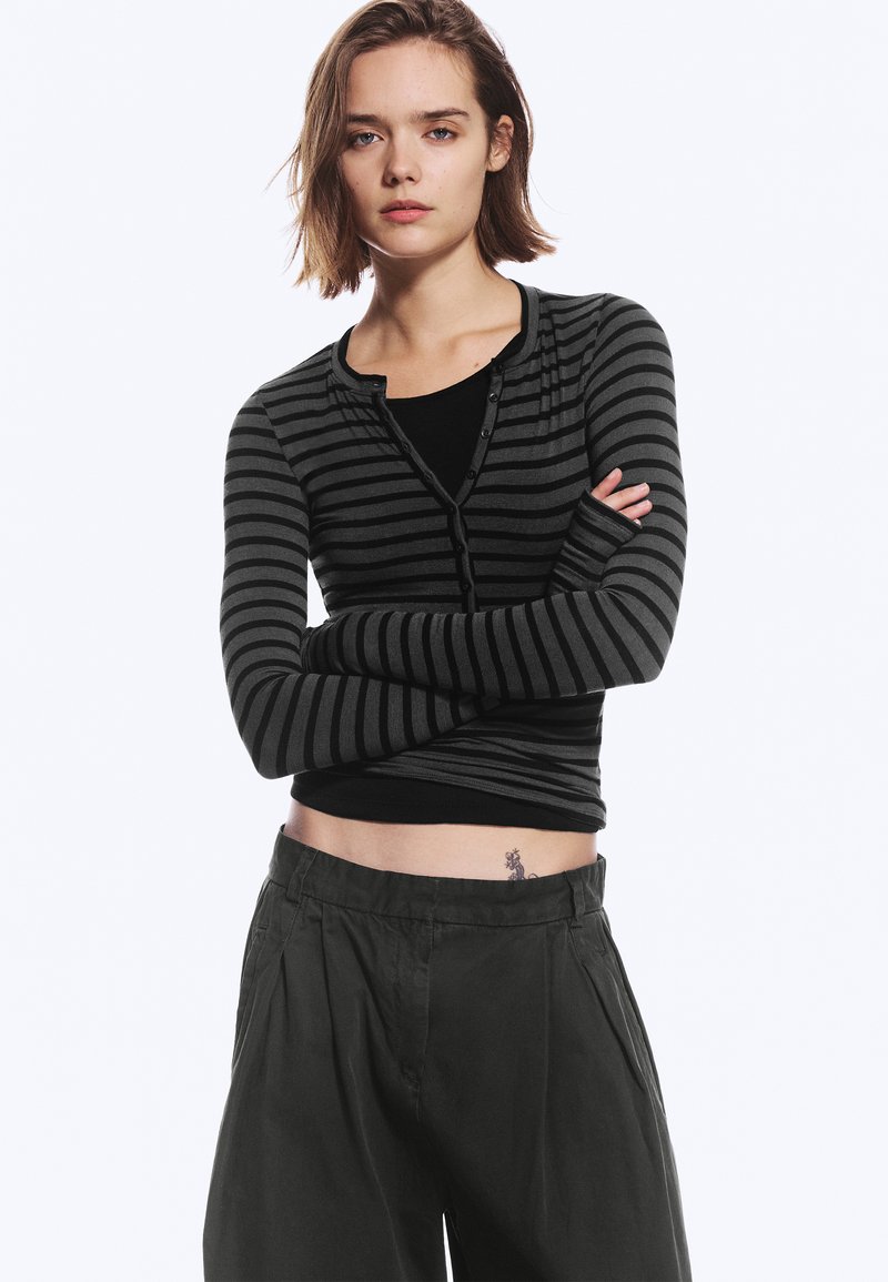 Une femme pose les bras croisés, vêtue d’un cardigan à rayures grises et noires sur un haut noir et d’un pantalon ample et sombre, devant un fond uni.