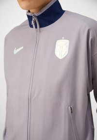 Nike tracksuit jacket in light gray, featuring a high navy collar, full zip, and logo patch with shield design on chest. Subtle texture.