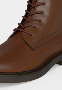 Bottine en cuir marron avec lacets ronds assortis et semelle en caoutchouc noire montrant des surpiqûres détaillées sur le côté et la zone des orteils.