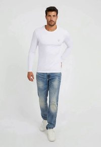 Camisa branca de manga longa com um pequeno logotipo, combinada com jeans desgastados de tom azul claro e sapatilhas brancas. Design simples e casual.