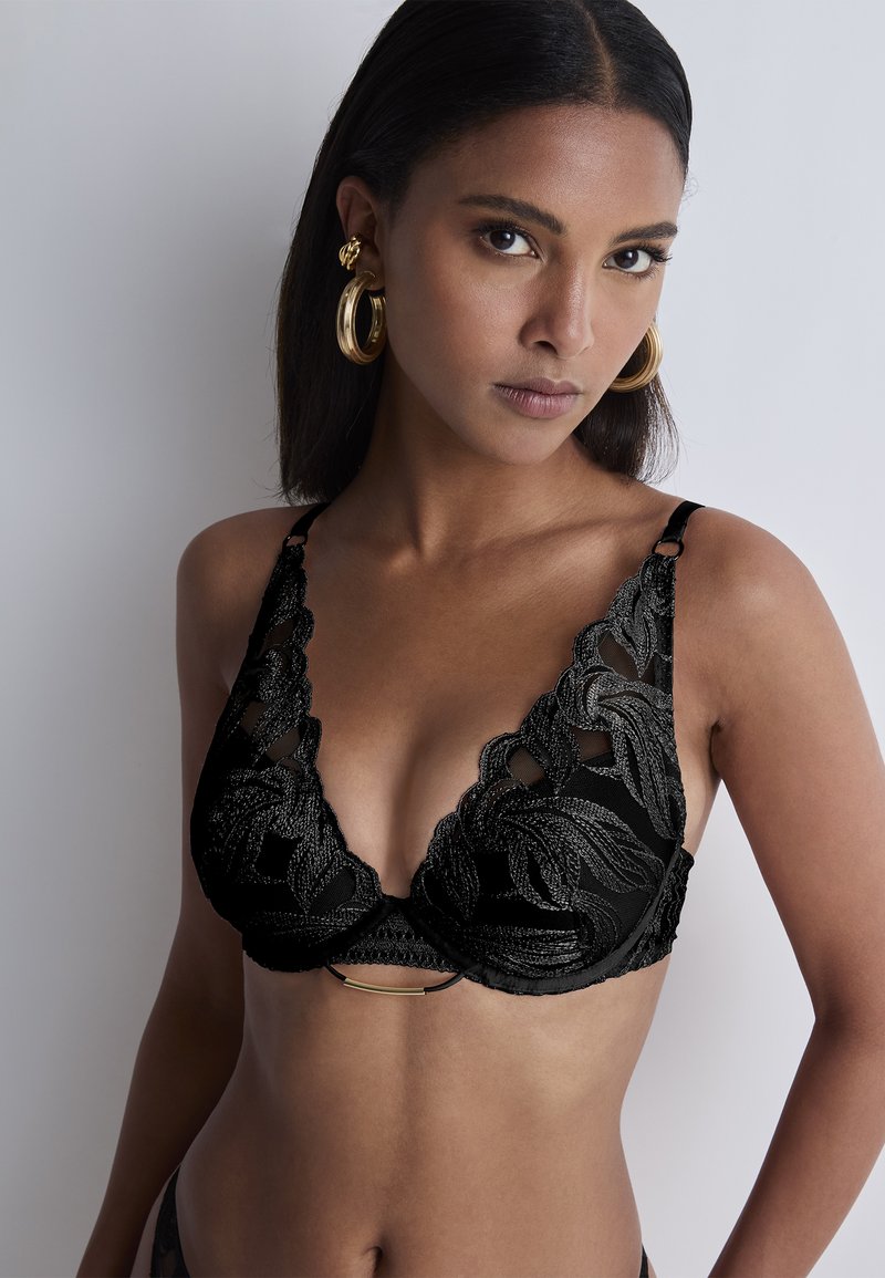 Soutien-gorge en dentelle noire avec un design en V profond, orné de motifs en forme de feuilles et d'un accent métallique au centre, avec des bretelles ajustables.