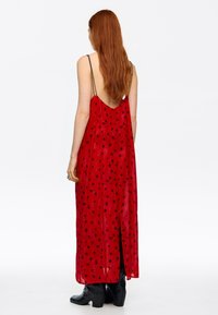 Kobieta o długich czerwonych włosach, ubrana w czerwoną sukienkę bez pleców w czarne kropki oraz czarne kozaki na obcasie, stoi na białym tle.