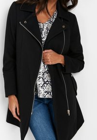 Černý kabát se strukturovaným designem, předním zipem a bočními kapsami. Texturovaná látka s ostrým límcem. Kombinován s vzorovaným topem a džíny.