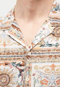 Πουκάμισο με γιακά και κουμπιά, διακοσμημένο με μοτίβα ήλιου, λουλουδιών και περίπλοκων σπειροειδών σχεδίων σε αποχρώσεις πορτοκαλί, μπλε και μπεζ.