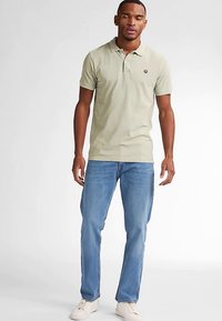 Hellgrünes Poloshirt mit kurzen Ärmeln und Kragen, kombiniert mit blauen Jeans aus Denim. Weiße Turnschuhe vervollständigen das Outfit und schaffen einen lässigen Look.