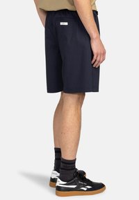 Pantaloni corti blu navy realizzati in tessuto leggero, con una tasca posteriore e un branding discreto, indossati con scarpe nere e calze a fantasia.