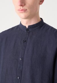 Marineblå linsekjorte med båndkrage, som har en frontplacket med knapper og et teksturert stoff. Nærbilde fremhever knappene og kragedesignet.