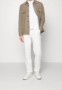 Bruin overhemd met knopen en twee borstzakken boven een wit T-shirt, gecombineerd met witte broek en lichtgrijze sneakers. Gladde stof, casual pasvorm.