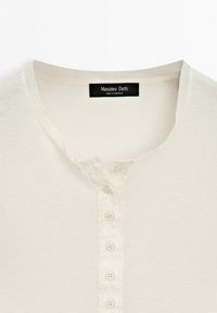 Chemise en maille couleur crème avec cinq petits boutons sur le devant et une étiquette noire portant l'inscription « Massimo Dutti Made in Morocco » à l'intérieur du col.