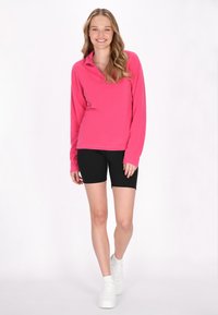 Pullover rosa in fleece a maniche lunghe con colletto con zip, abbinato a pantaloni shorts neri da ciclismo e sneakers bianche, su sfondo bianco semplice.