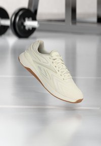 Αθλητικό παπούτσι σε ανοιχτό μπεζ χρώμα με σόλα από καουτσούκ, αιωρούμενο πάνω από το δάπεδο του γυμναστηρίου, με μια θολή αλτήρα και πάγκο στο φόντο.