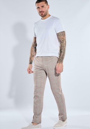Man with tattooed arms wearing a white t-shirt, beige checkered pants, and white slip-on shoes, standing against a plain light background.