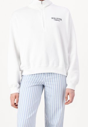 Weißer Zip-Up-Hoodie mit hohem Kragen und gerippten Bündchen, mit einem kleinen schwarzen Logo "Hollister California" auf der Brust. Kombiniert mit gestreiften blauen Hosen.