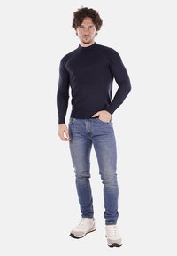 Maglione a dolcevita blu navy realizzato in tessuto morbido, design aderente, indossato con jeans azzurri e scarpe da ginnastica bianche, in piedi su uno sfondo bianco.