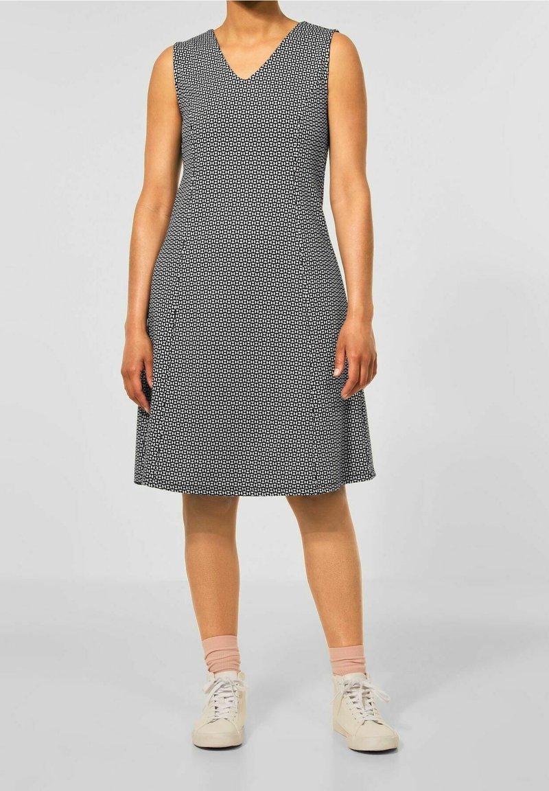 Femme portant une robe sans manches noire et blanche à motifs de longueur genou, avec des chaussettes roses et des baskets blanches sur un fond uni.