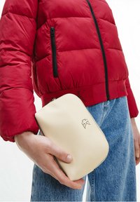 Pochette in pelle crema con logo dorato, tenuta in mano. Accompagnata da un giubbino imbottito rosso e jeans in denim blu. Compatta con una superficie liscia.