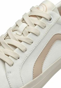 Baskets blanches en toile avec des accents beiges, dotées de lacets étroits, d'une surface texturée et d'une semelle en caoutchouc lisse.