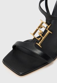 Μαύρο δερμάτινο πέδιλο με ψηλό τακούνι και τετράγωνη μύτη, με χρυσή διακοσμητική λεπτομέρεια λογότυπου και λεπτό λουράκι στον αστράγαλο. Ομαλή υφή και λιτό σχέδιο.