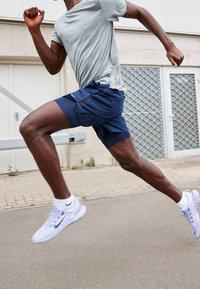 Hombre atlético corriendo en el pavimento, vestido con una camiseta de manga corta gris claro y pantalones cortos azul marino, con zapatillas de running blancas y piernas musculosas destacadas.