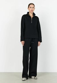 Svart zip-up sweatshirt med hög krage, vida ärmar och matchande löst sittande byxor. Modellen bär svart och vitt randiga skor.