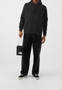 Sweatshirt noir à capuche avec une poche avant, associé à un jean noir ample et des baskets Vans noires. Tient un petit sac noir avec un logo.