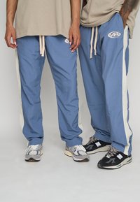Blå lätta byxor med krämfärgade sidosträck och en broderad logotyp. Matchat med grå och svarta sneakers, med texturerade material.