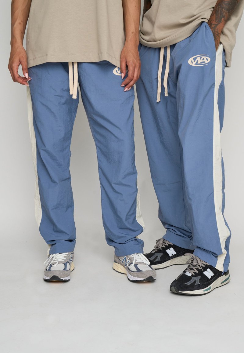 Blå lätta byxor med krämfärgade sidosträck och en broderad logotyp. Matchat med grå och svarta sneakers, med texturerade material.