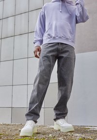 Osoba stojąca na zewnątrz, ubrana w lawendowy hoodie, szare dżinsy i białe trampki, na żwirowym podłożu z tłem z kafelkowej ściany.