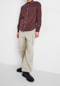 Camisa de pana marrón con dos bolsillos en el pecho, combinada con pantalones cargo de pierna ancha color beige claro y botas negras hasta el tobillo. Fondo sencillo.