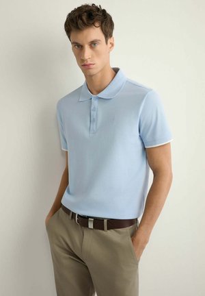 Młody mężczyzna w jasnoniebieskiej koszulce polo i beżowych spodniach z brązowym paskiem, stojący z rękami w kieszeniach na tle jednolitej ściany.