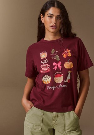 Woman wearing a maroon t-shirt with fall-themed illustrations including a candle, cup, leaves, pumpkin, cinnamon roll, basket, dog, and "Cosy Season" text.