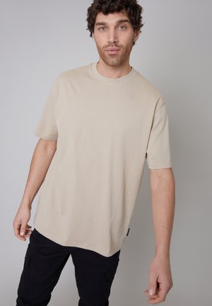 Beige cotton t-shirt with short sleeves, round neck, relaxed fit, and subtle hem detailing, paired with black cargo pants.