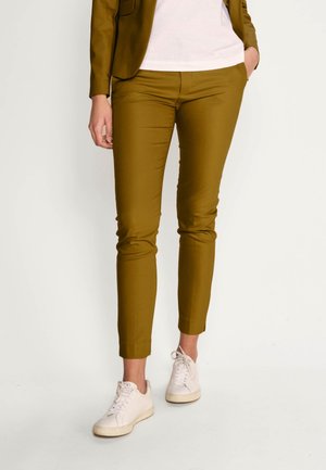 Person iført smalle bukser i sennepsfarve, hvide sneakers og en matchende sennepsfarvet blazer med en hvid skjorte, stående med den ene hånd i lommen.
